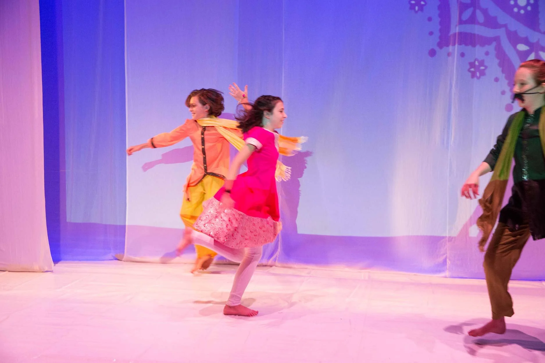 Meninas dançando no palco com figurinos coloridos e fundo decorado com temas natalinos.