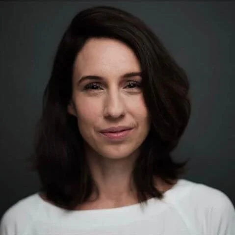 Retrato de uma mulher com cabelo castanho escuro, usando uma blusa branca, com fundo escuro.