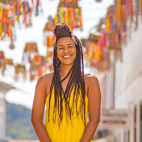 Mulher sorridente de cabelos trançados usando vestido amarelo, ao ar livre, com bandeirinhas coloridas decorativas no fundo.