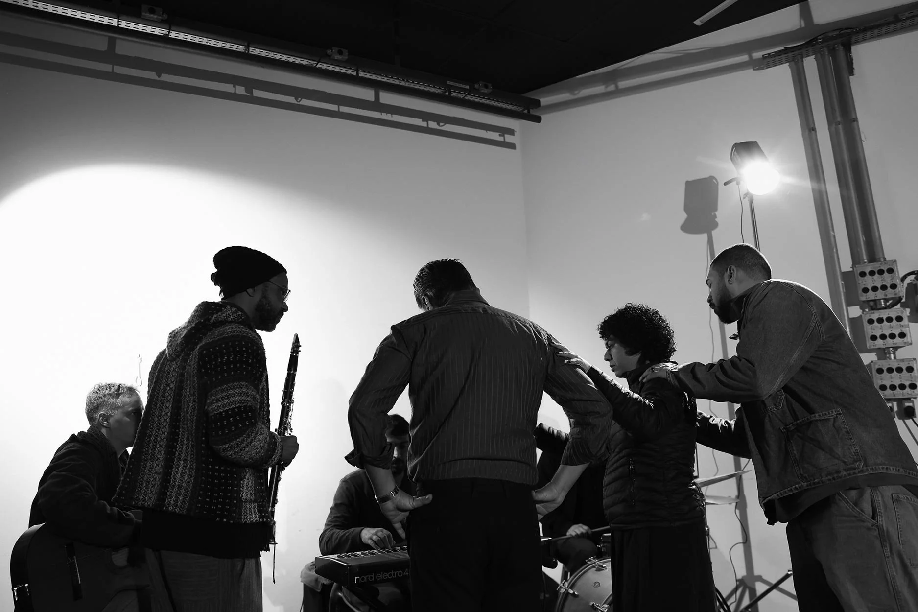 Grupo de pessoas tocando instrumentos musicais, incluindo um clarinete e um teclado, em um palco com iluminação de estúdio