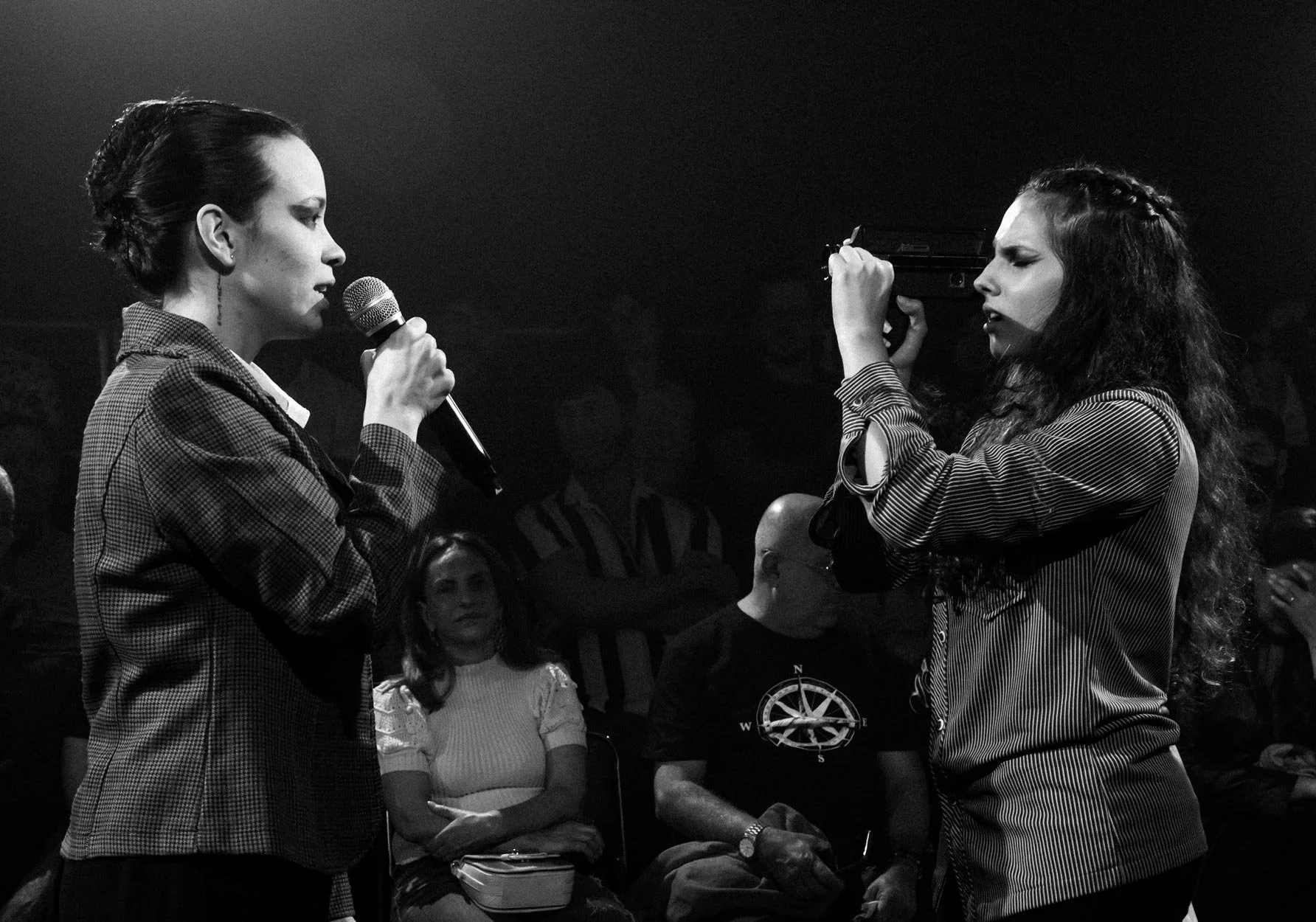 Duas mulheres em uma conversa cerrada no palco, uma segurando um microfone e a outra uma arma, em um ambiente escuro com várias pessoas ao fundo observando.
