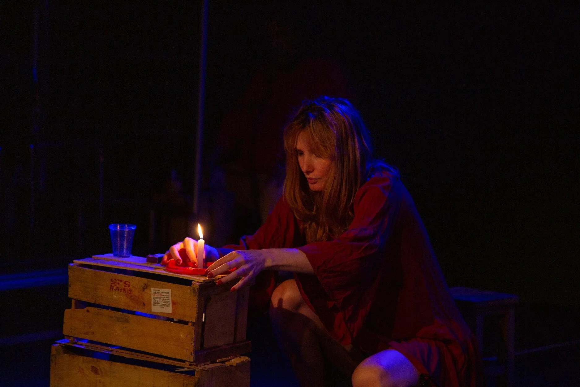 Mulher com cabelo castanho avermelhado sentada em um palco escuro, usando um vestido vermelho, acendendo uma vela em uma caixa de madeira com um fundo negro.