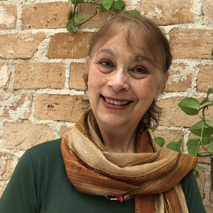 Mulher sorrindo com cachecol laranja, de cabelo castanho, em frente a uma parede de tijolos, com plantas verdes ao fundo.