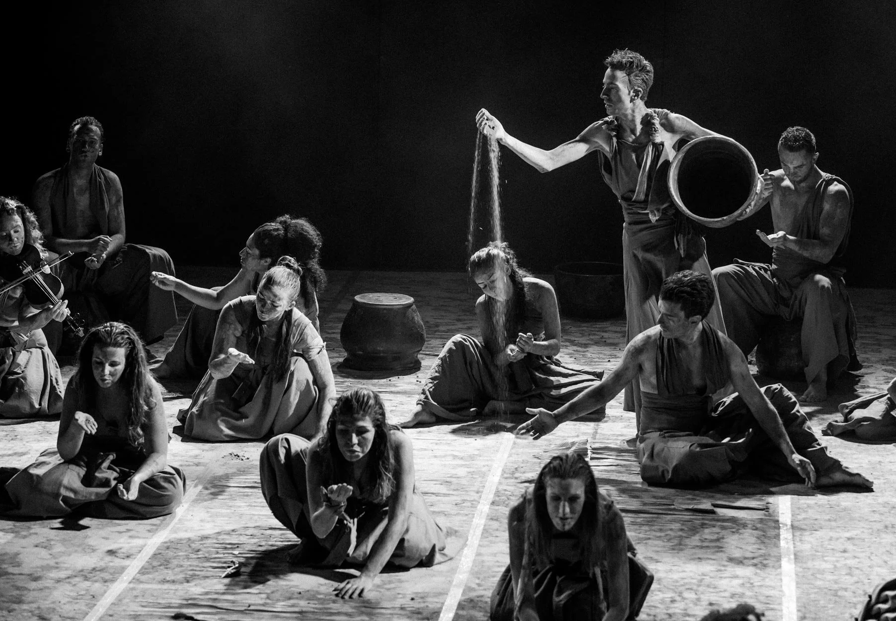 Cena de uma apresentação teatral em preto e branco com várias pessoas no palco, algumas sentadas, outras de joelhos, uma pessoa de pé segurando um recipiente e jogando areia, todas com roupas soltas e expressionistas, sugerindo uma obra dramática ou 