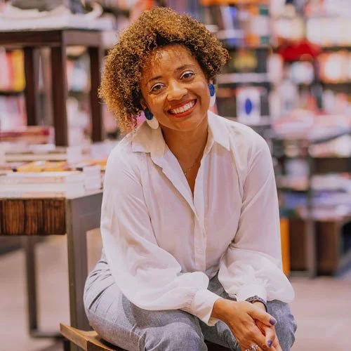 Mulher sorrindo, com cabelo cacheado, usando blusa branca, sentada em uma loja de livros.