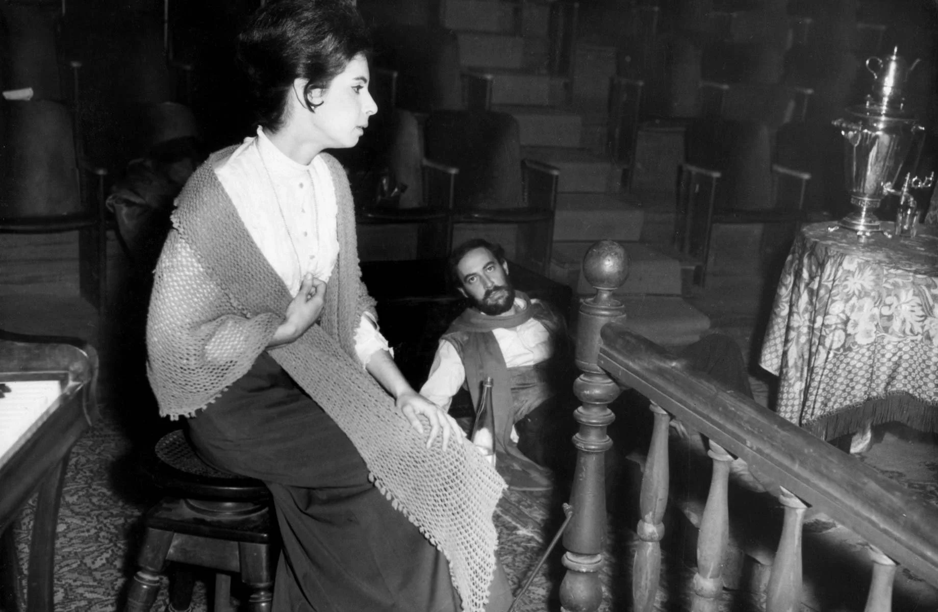 Cena de uma mulher com expressão séria, sentada em uma cadeira, vestindo roupas simples com um suéter grosso, ao lado de uma escadaria de madeira, observando um homem de barba, deitado no chão, com roupas antigas, em um ambiente escuro, possivelmente