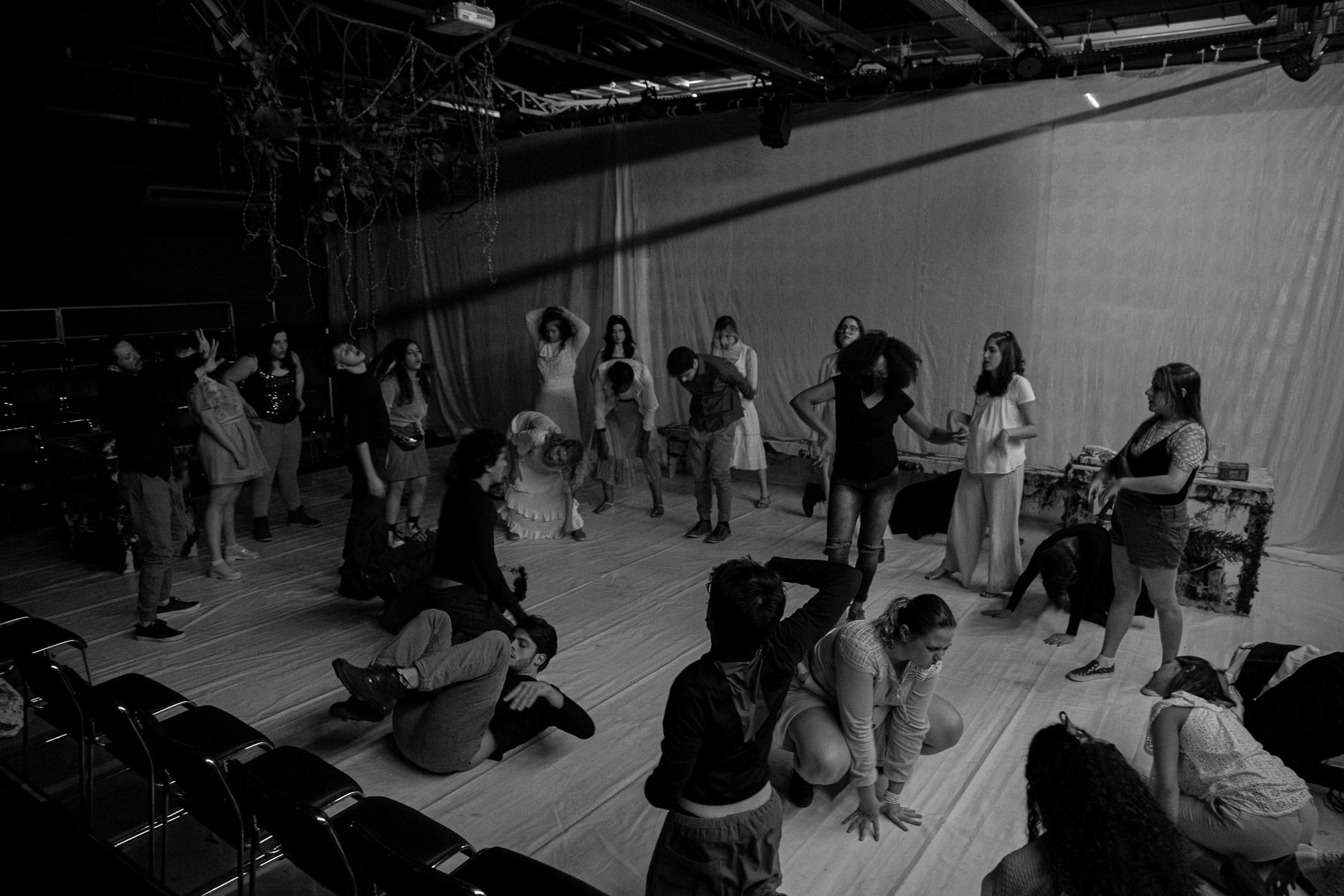 Grupo de pessoas ensaiando ou participando de uma peça de teatro, alguns estão deitados no chão, outros estão em pé, em um espaço que parece ser um teatro ou estúdio de gravação, com cortinas ao fundo e equipamentos de iluminação pendurados no teto.