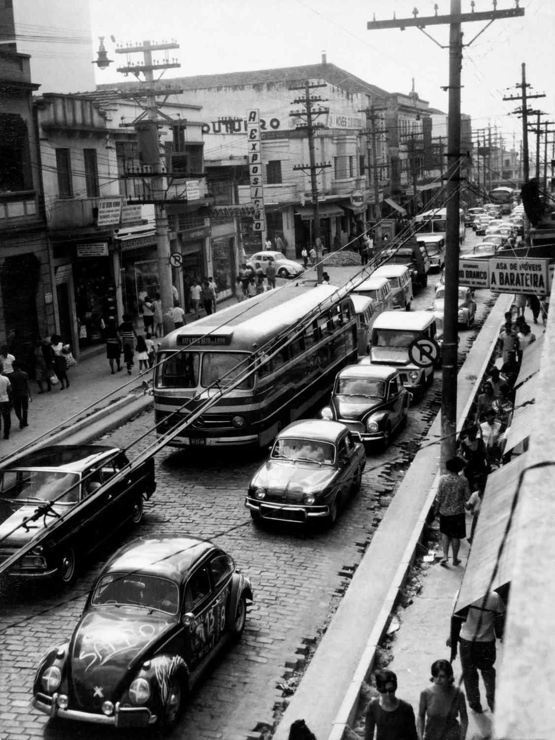 Imagem em preto e branco de uma rua movimentada com carros de época e linhas de ônibus, com pessoas caminhando na calçada e prédios ao fundo, incluindo uma loja de correios e sinais de trânsito.