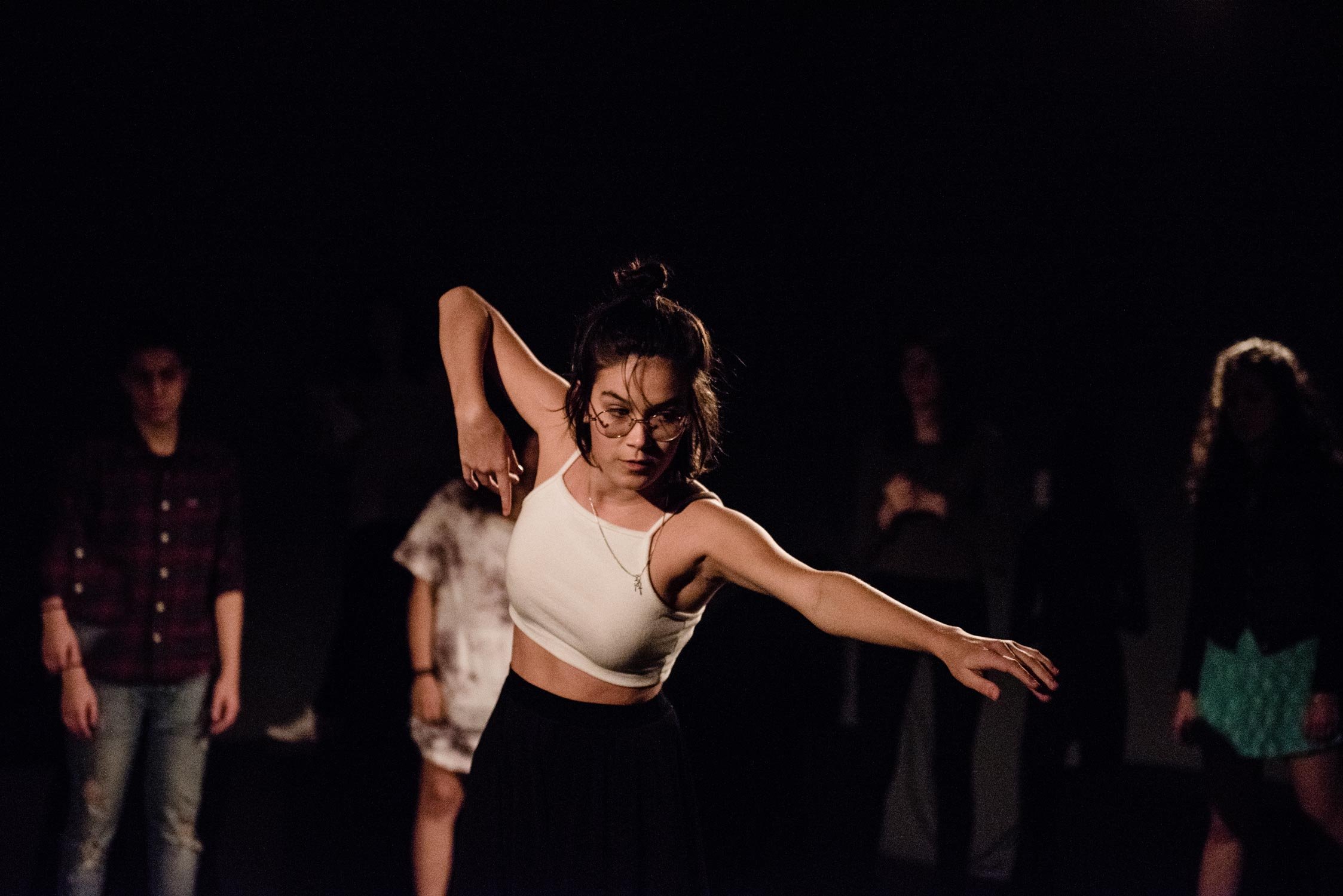 Juventude dançando em uma sala escura com foco na mulher de óculos ao centro, vestindo blusa branca e saia preta.