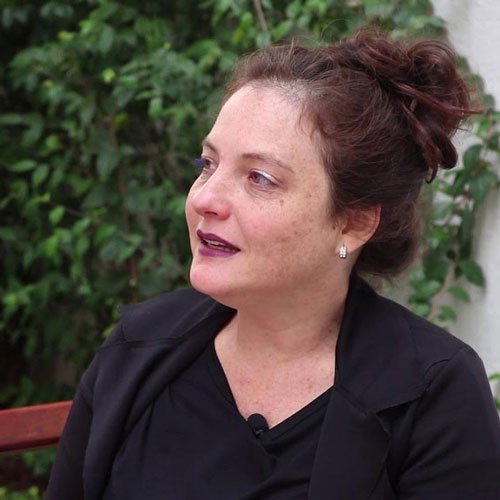 Mulher com cabelo castanho preso, usando blusa preta, sentada ao ar livre com vegetação ao fundo.