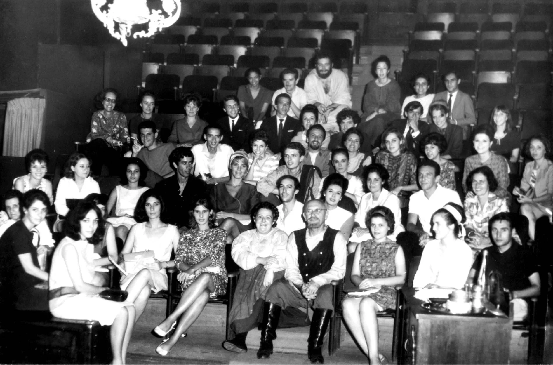 Fotografia em preto e branco de um grupo grande de pessoas em uma sala de teatro, algumas sentadas no palco e outras nas cadeiras do público.