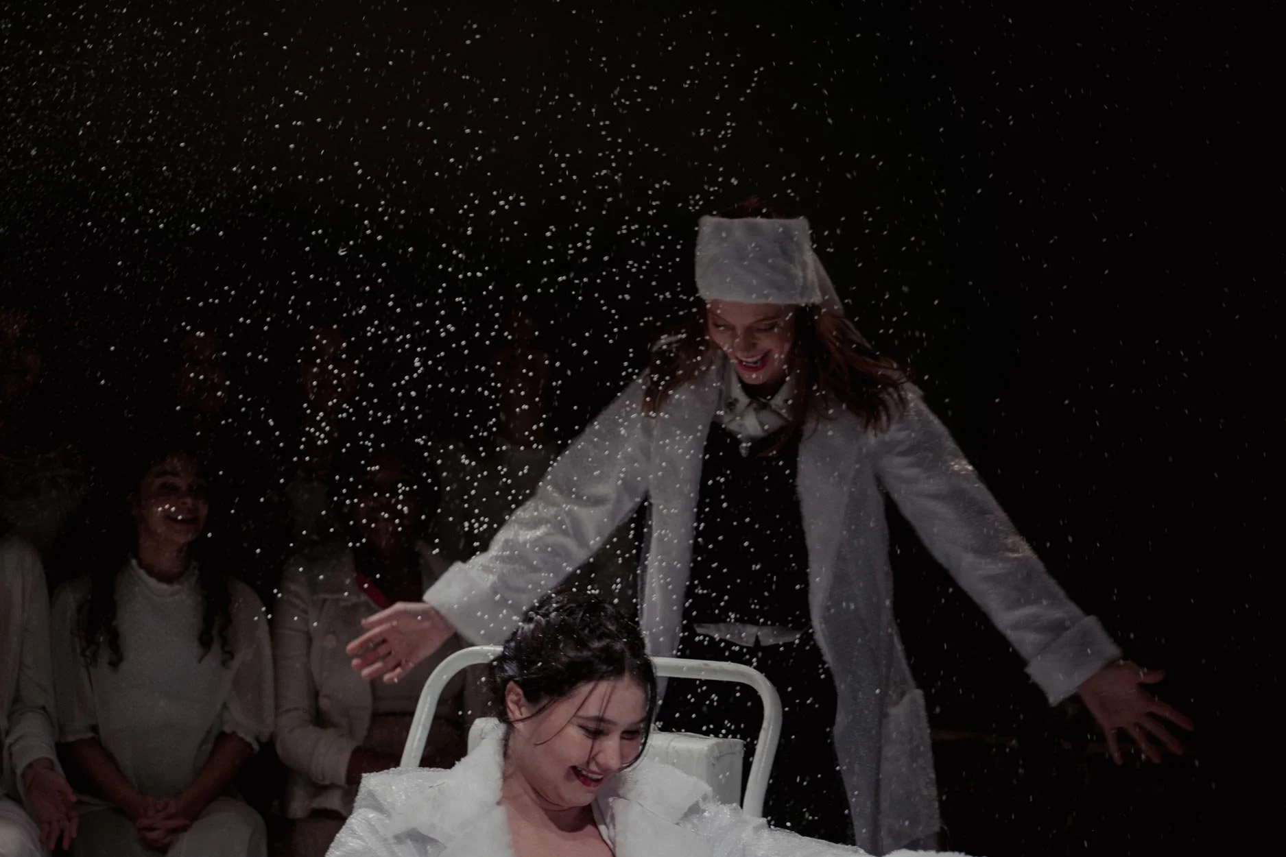 Mulher vestida de branco sorrindo na cama de hospital enquanto pessoas ao fundo riem, com uma mulher de avental e touca na frente, durante parto ou procedimento médico