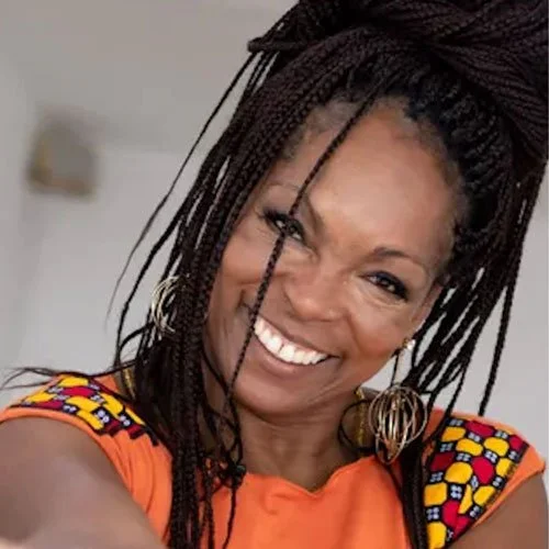 Mulher sorridente com dreadlocks, usando uma blusa laranja com detalhes coloridos e brincos grandes de argola.