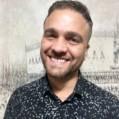 Homem sorrindo, de camisa com estampa de bolinhas, em frente a fundo desfocado.