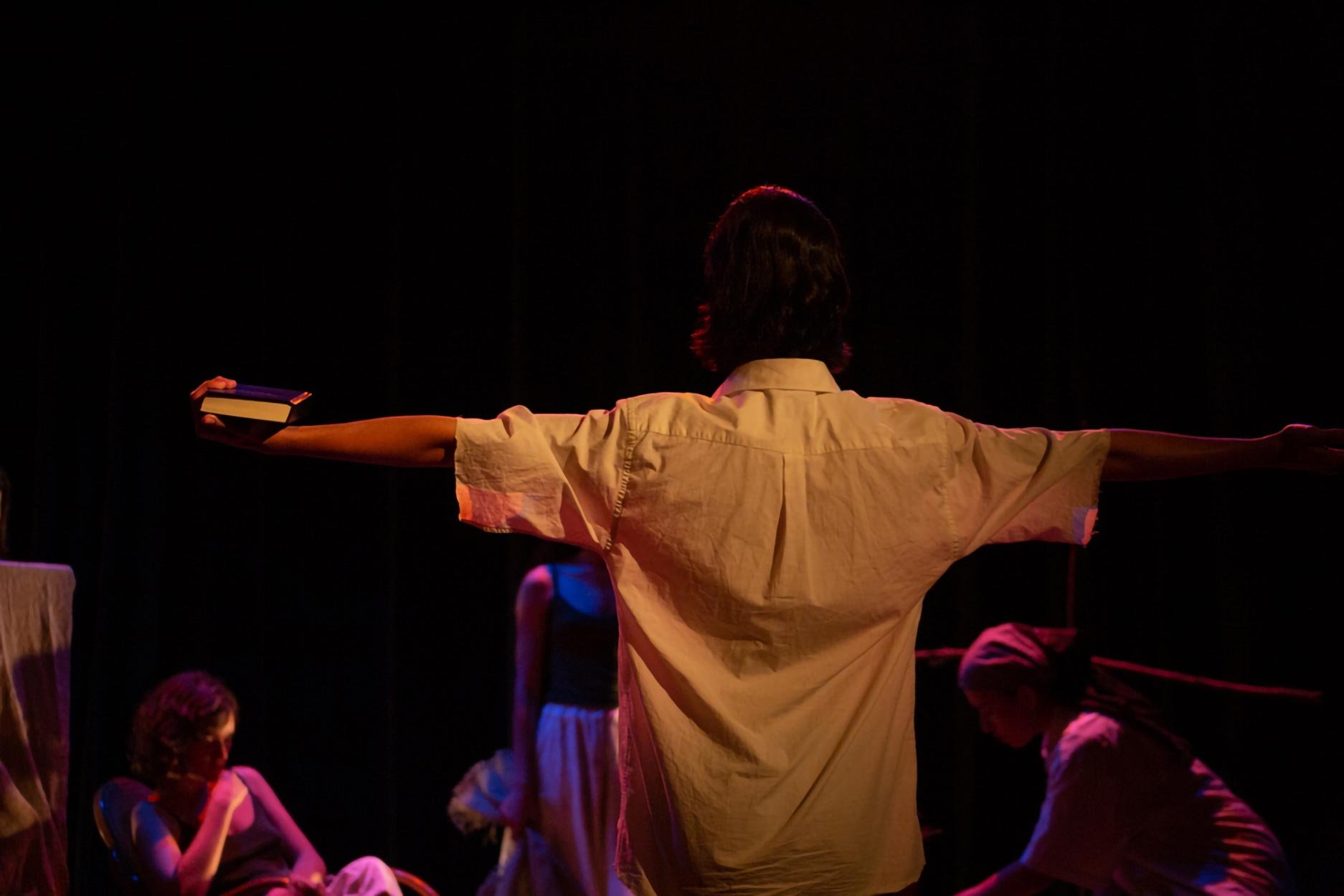 Pessoa de costas com braços abertos em um palco, com outras pessoas ao redor, sob iluminação dramática.