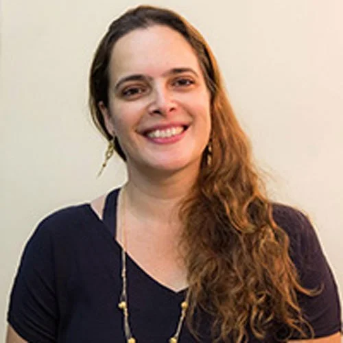 Mulher sorrindo com cabelo cacheado, usando camiseta azul escura e brincos dourados