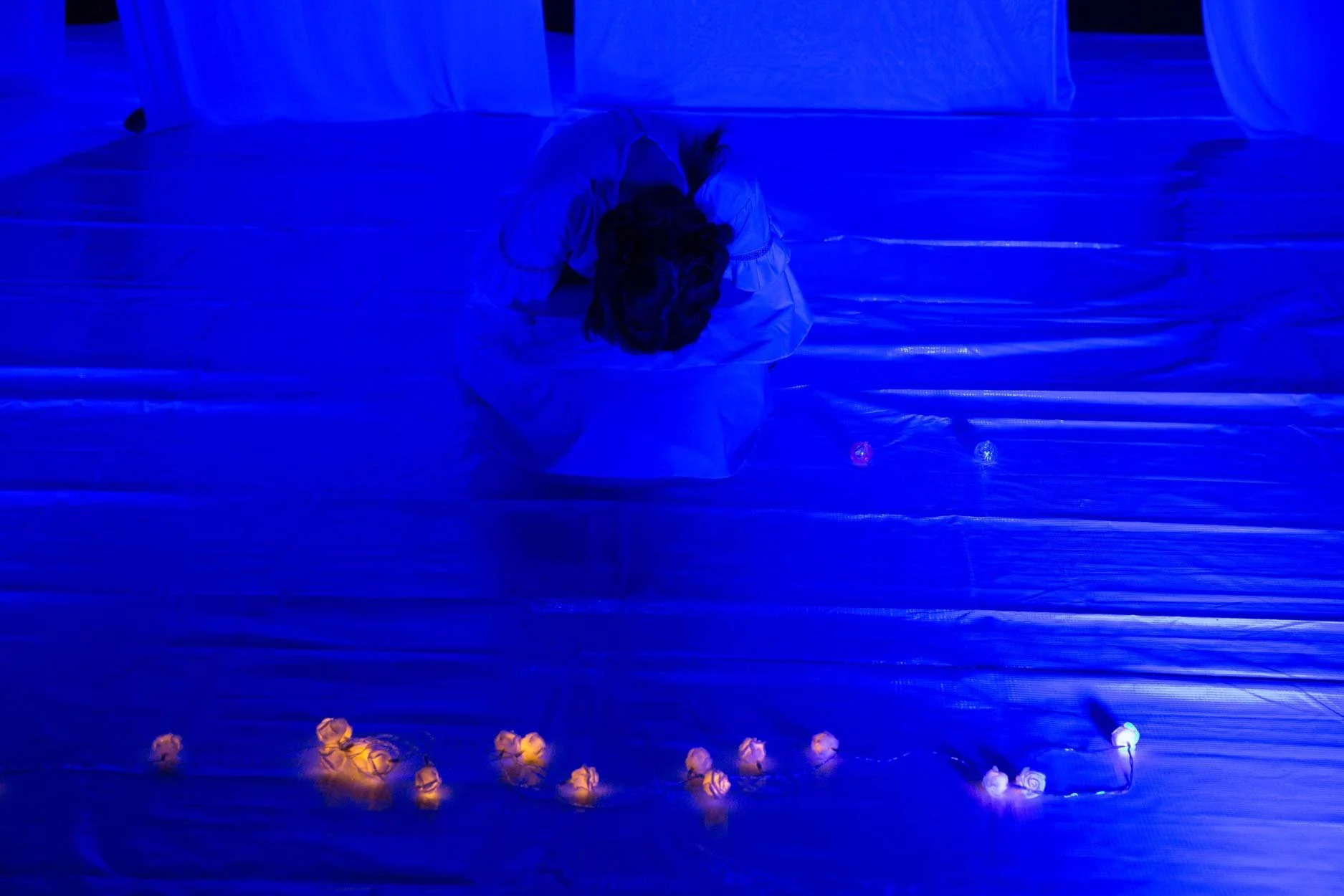 Pessoa deitada no chão sob luz azul, com pequenos objetos de luz coloridos espalhados ao redor.