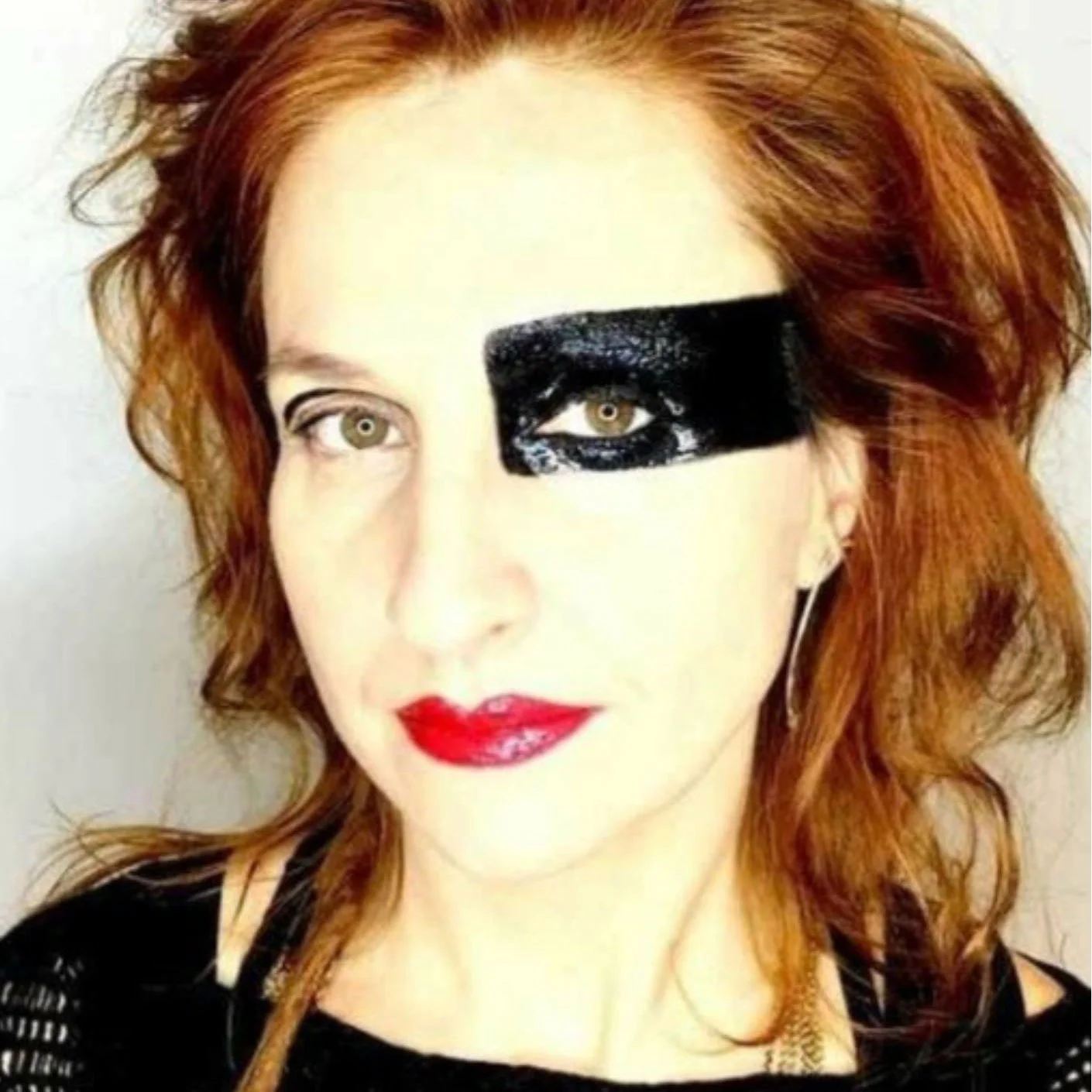 Mulher com cabelo ruivo e maquiagem marcante, usando uma faixa preta ao redor do olho direito, criando um efeito artístico, com batom vermelho, vestindo roupa preta de malha.
