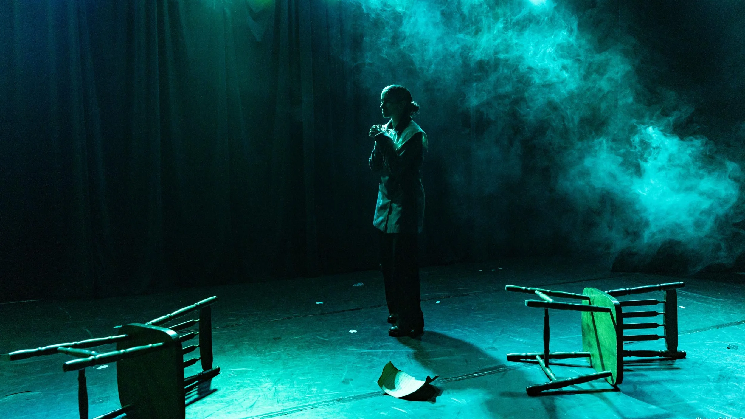 Pessoa de pé no palco, cercada de fumaça, com cadeiras de palha derrubadas ao redor, em um ambiente escuro com iluminação teatral