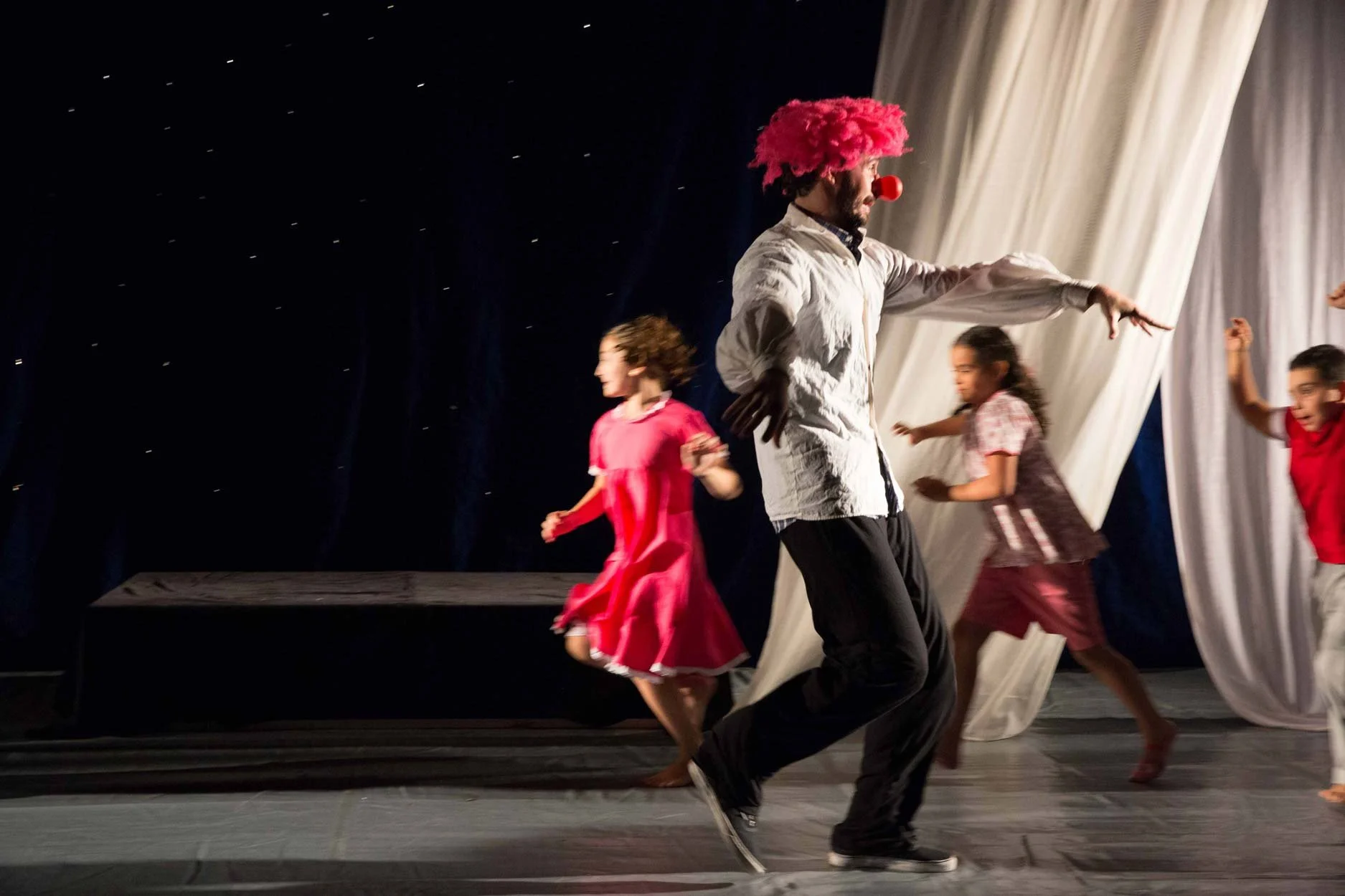 Homem fantasiado de palhaço com peruca rosa, jaqueta branca, calça preta e nariz vermelho, dançando com crianças em palco com cortina branca ao fundo.