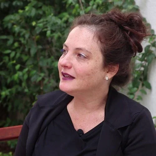 Mulher com cabelo castanho preso, usando brincos de pérola, vestindo roupa preta, sentada ao ar livre com vegetação ao fundo.