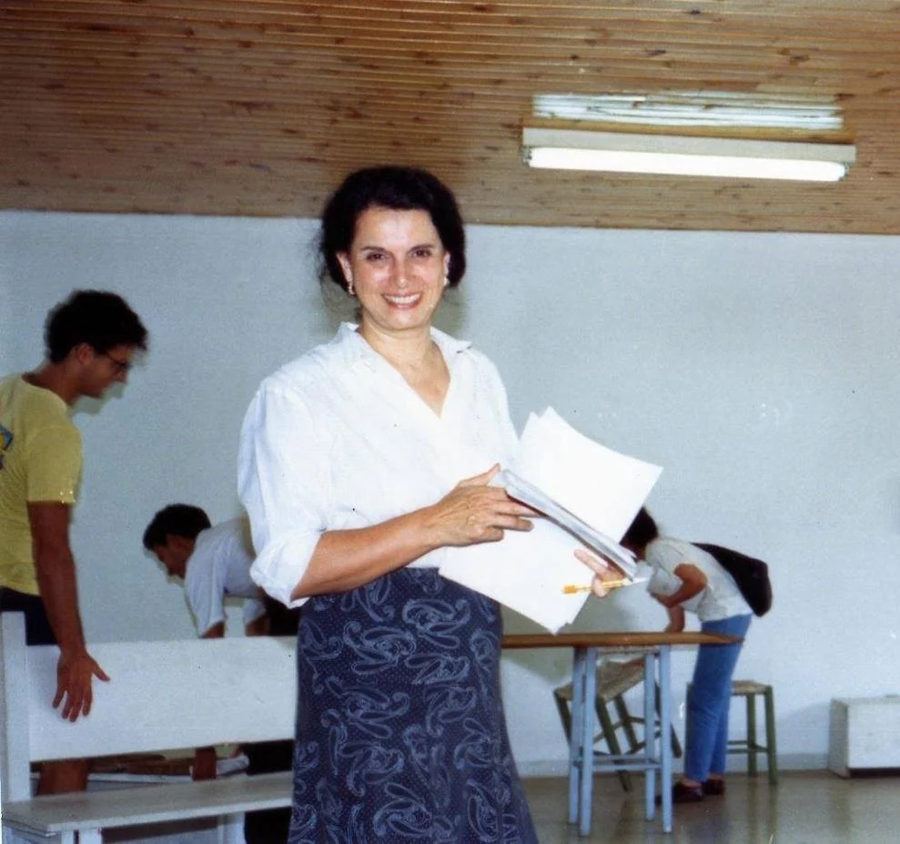 Mulher sorridente segurando papéis, em sala com pessoas ao fundo e teto de madeira