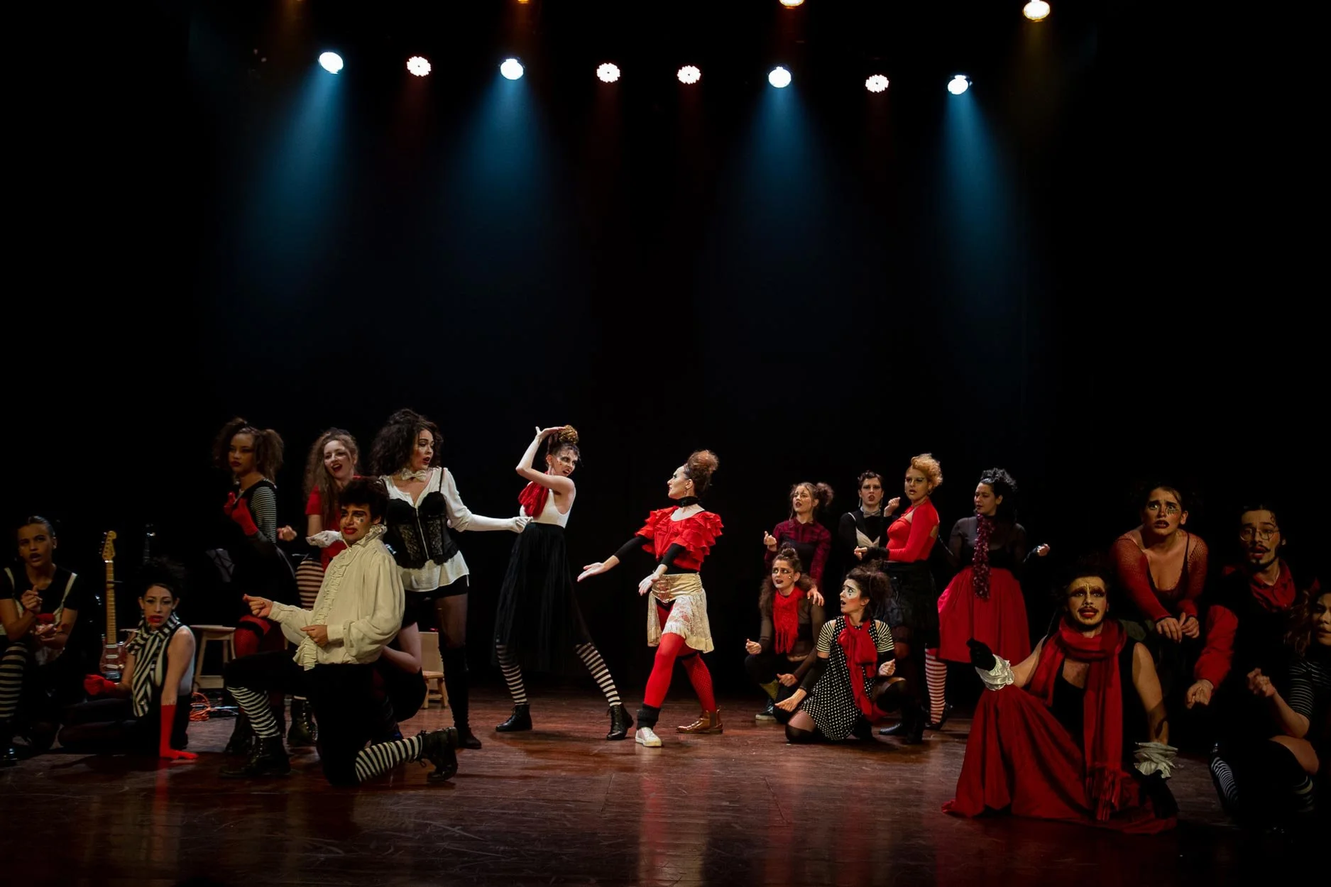 Grupo de atores em cena de teatro, usando figurinos coloridos em um palco com iluminação dramática.