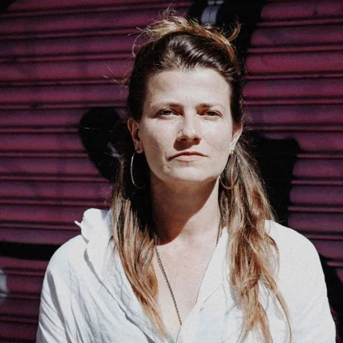 Mulher com cabelo castanho claro, usando brincos de argola e roupa branca, em frente a uma porta vermelha de aço.