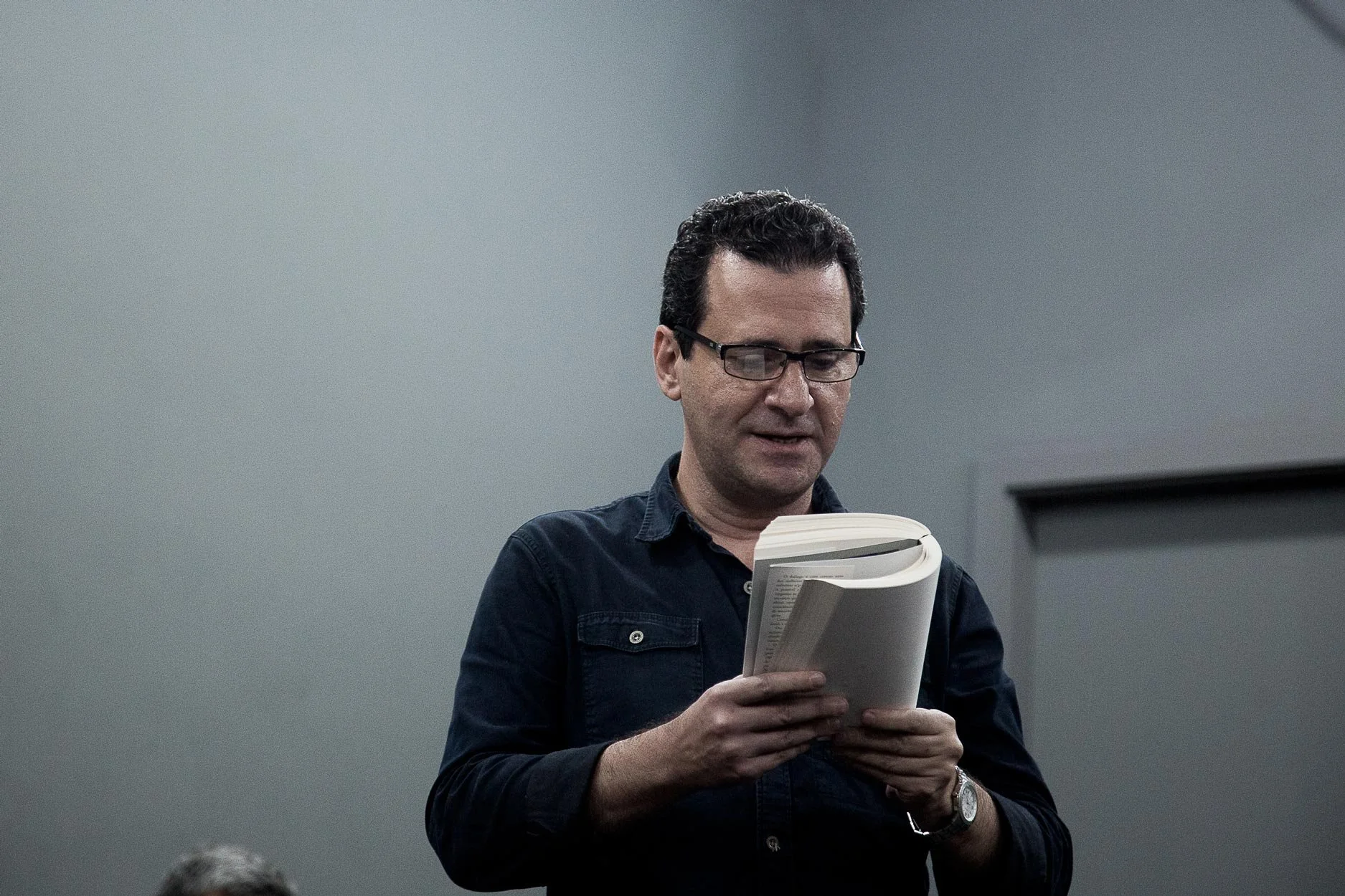 Homem com óculos lendo um livro ou caderno em um ambiente interno com paredes cinzas.