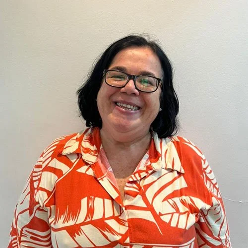 Mulher sorridente com cabelo preto, óculos e camisa laranja e branca com padrão de folhas, posando contra uma parede cinza.