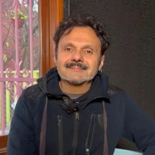 Homem sorridente com cabelo cacheado grisalho, usando jaqueta preta, sentado perto de uma janela com grades rosa e fundo escuro.