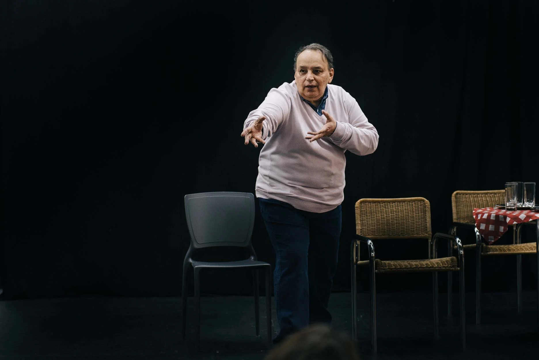 Homem fazendo uma apresentação ou discurso em um palco escuro, com cadeiras ao redor e uma mesa com uma toalha vermelha e branca, e copos d'água.