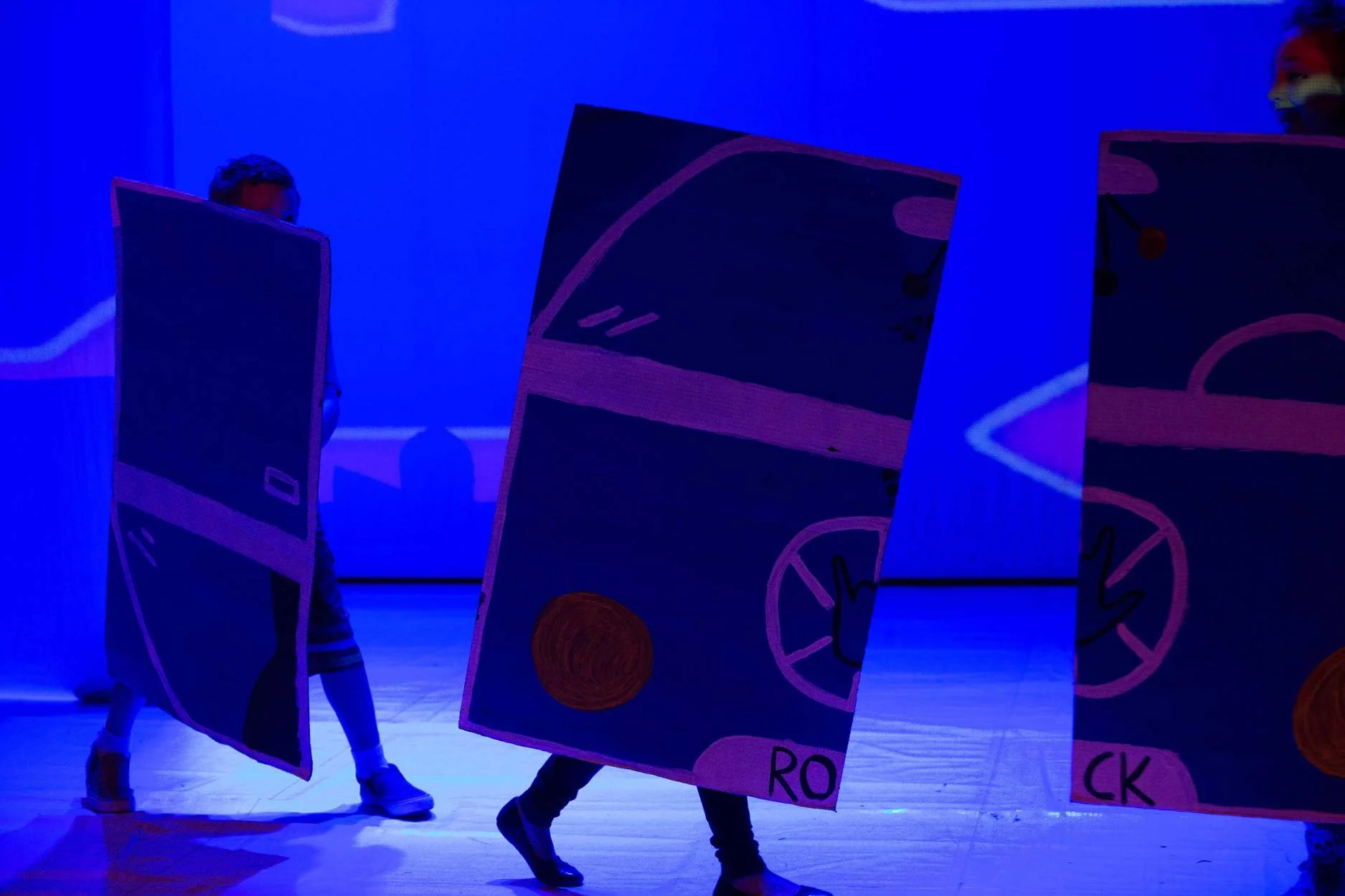 Crianças em uma apresentação de teatro com roupas e painéis que parecem de cartas de baralho, sob iluminação azul.