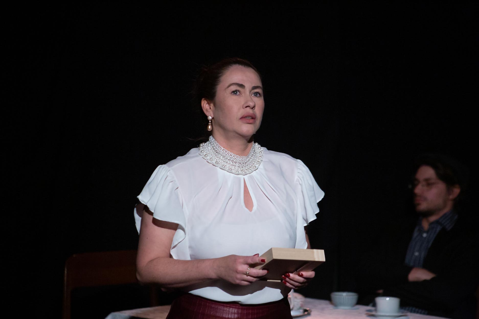 Mulher com roupas elegantes, brincos e colar de pérolas, segurando um livro, em cena dramática.