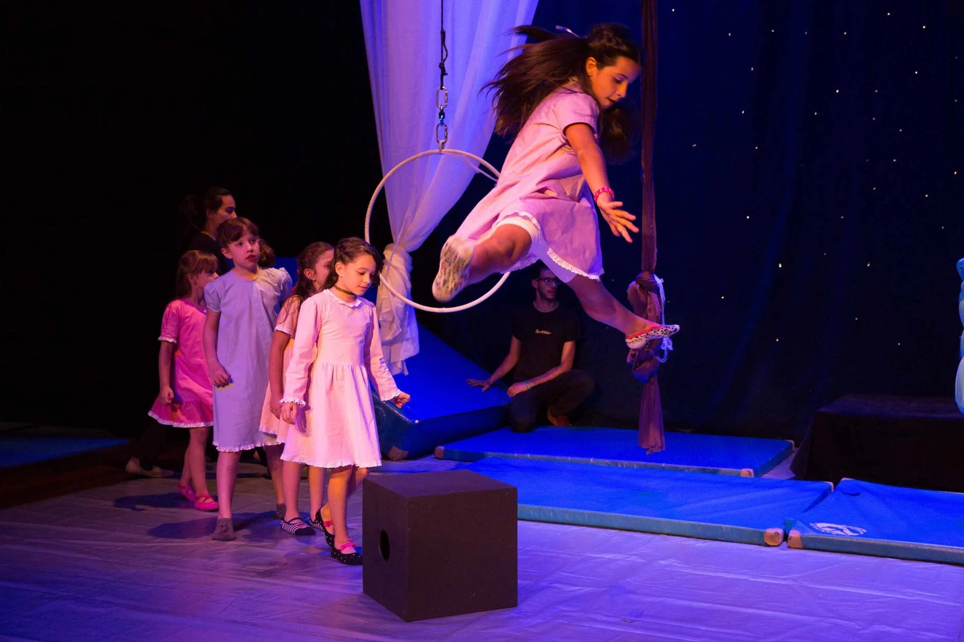 Crianças assistindo uma apresentação de circo, com uma menina pendurada em um trapézio e algumas outras em fila aguardando sua vez, tudo em um palco com iluminação azul escura.