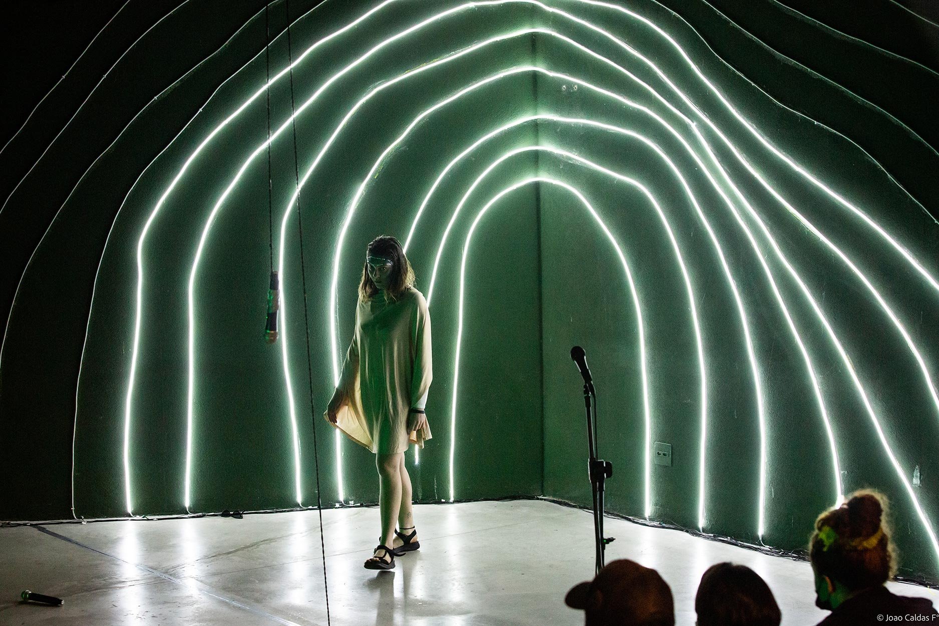 Mulher em palco diante de parede com luzes de LED verdes formando arcos, usando vestido claro e óculos de proteção, ao lado de microfone e fio de luz pendurado.