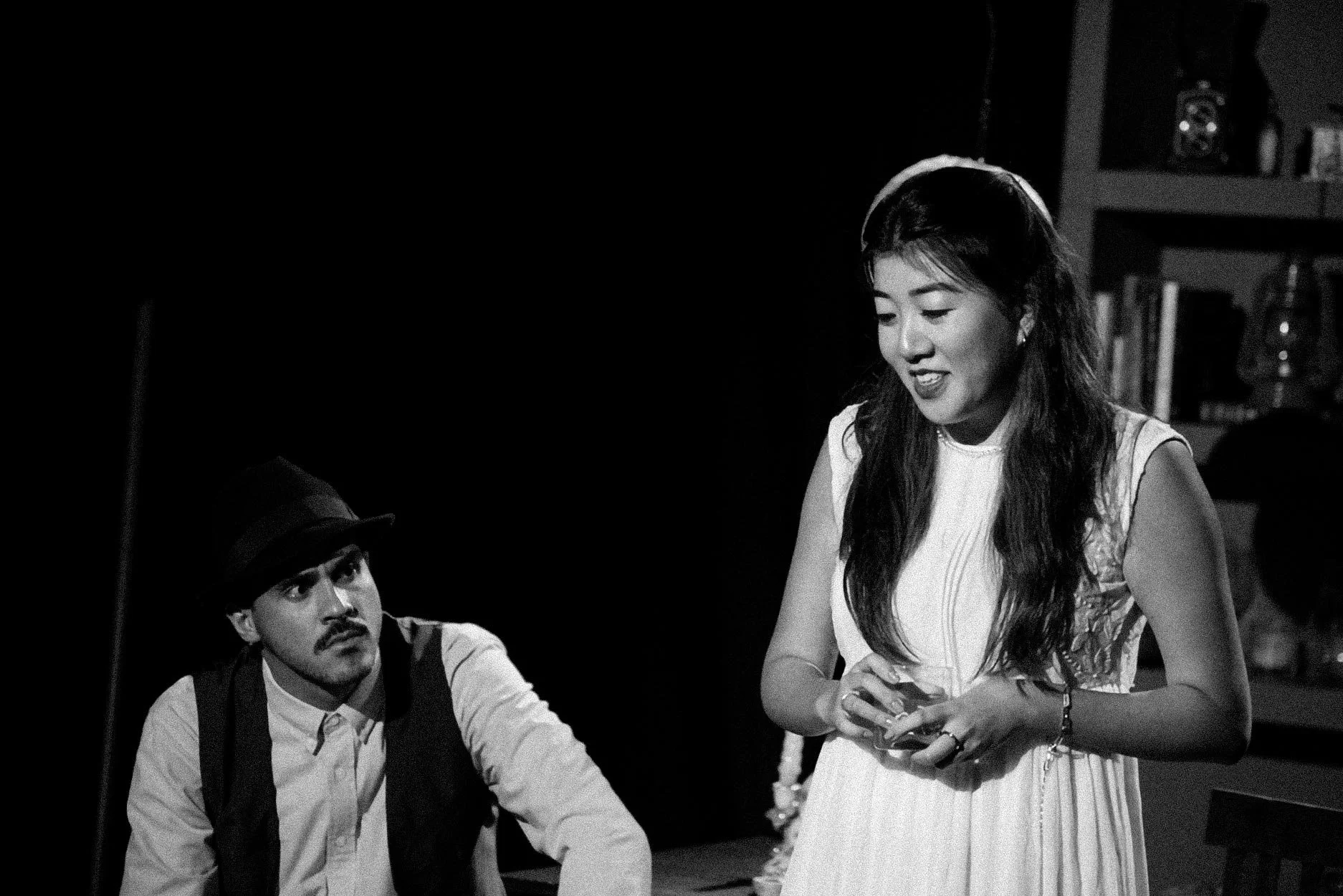 Imagem em preto e branco de uma mulher sorridente segurando um celular, ao lado de um homem com chapéu, ambos em um ambiente de sala com livros ao fundo.