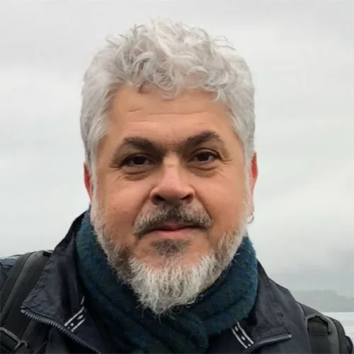 Homem de cabelo cinza e barba usando jaqueta e cachecol, em ambiente ao ar livre com céu nublado.