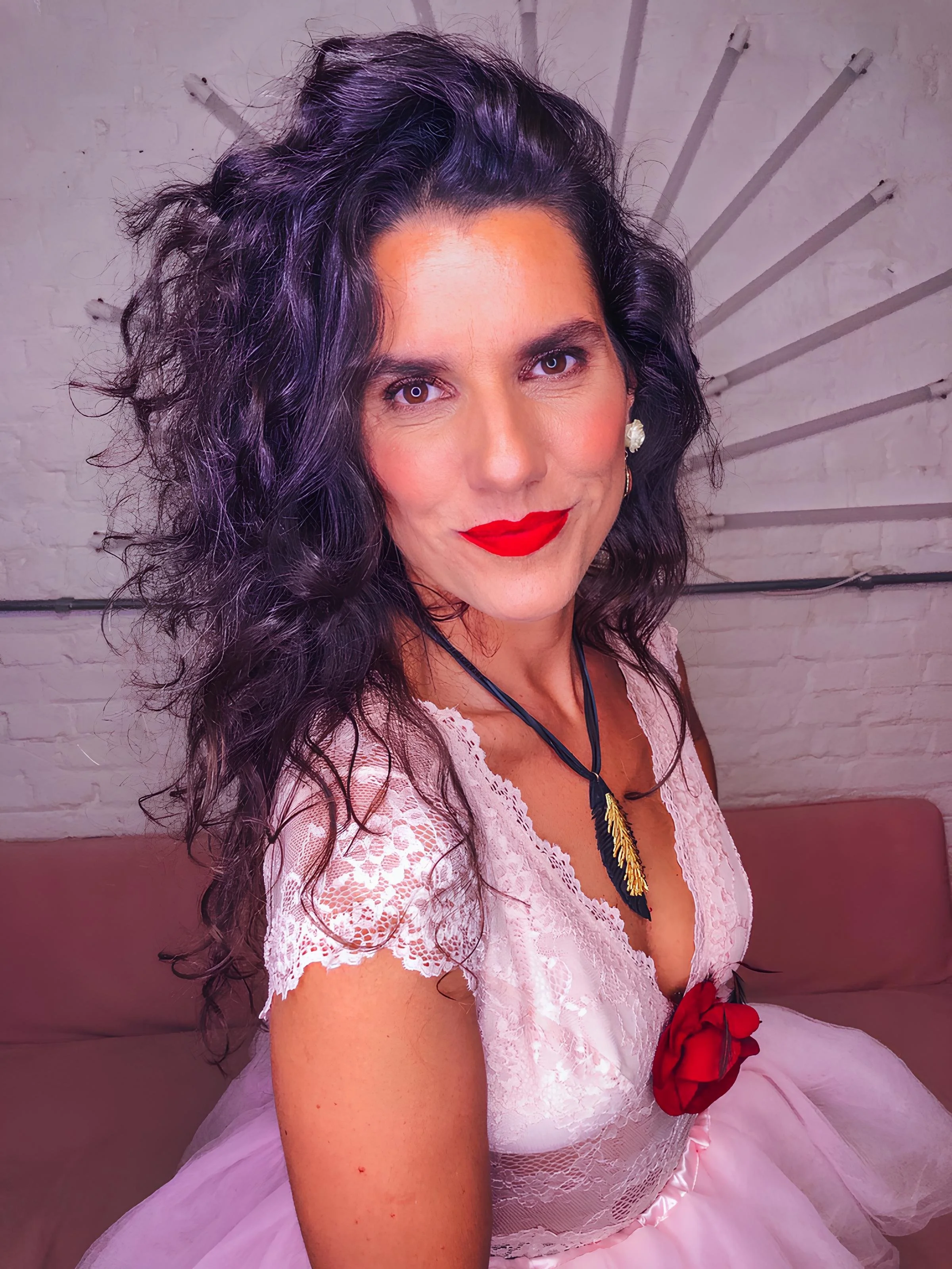 Mulher sorridente com os braços levantados, de cabelo cacheado, usando vestido marrom e colar. Ao fundo, decoração colorida e plantas.