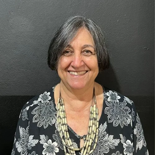 Mulher idosa sorridente com cabelo cinza, usando blusa com estampa de flores e colar de várias camadas, contra parede cinza.