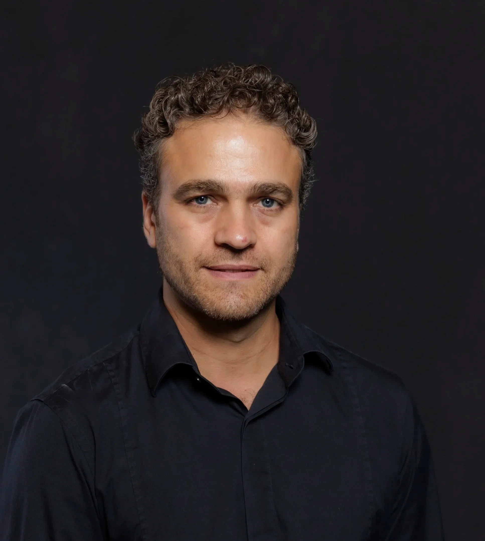 Homem sorridente com barba, cabelo castanho claro, usando camisa de botão com estampa de pontos brancos, em fundo com parede decorada com desenhos abstratos.