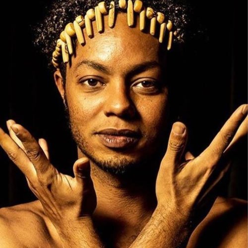 Pessoa negra com cabelo encaracolado usando presilhas de madeira formando uma coroa, fazendo um gesto com as mãos cruzadas na frente face]