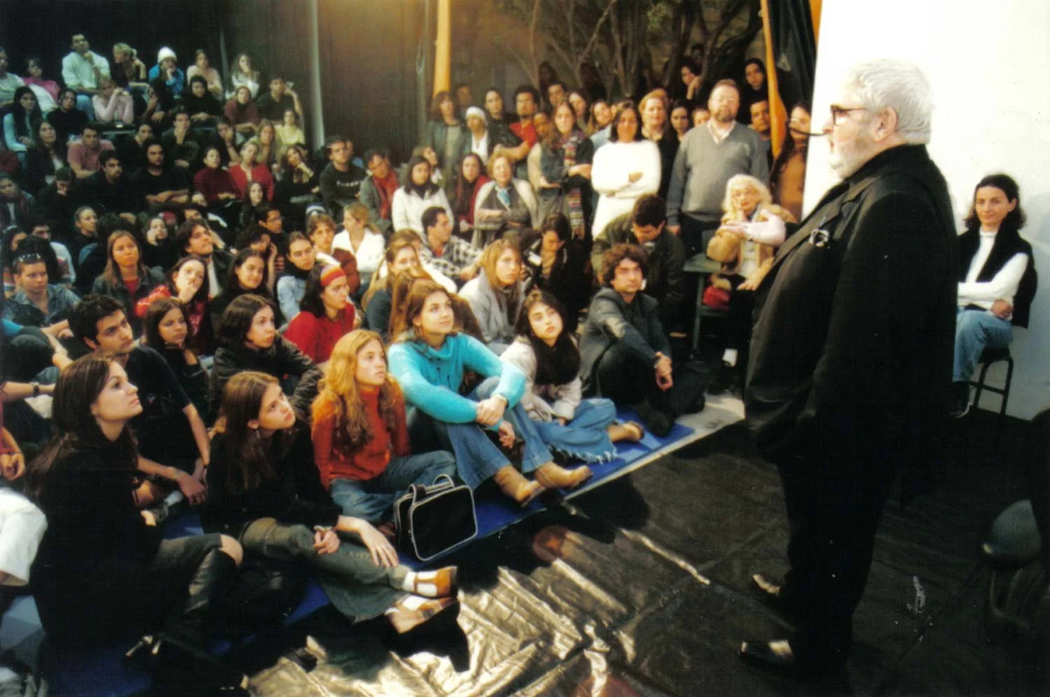 Homem de cabelos brancos e barba, vestindo roupas pretas, fala para uma plateia de jovens e adultos sentados e em pé, em um ambiente de palestra ou aula.