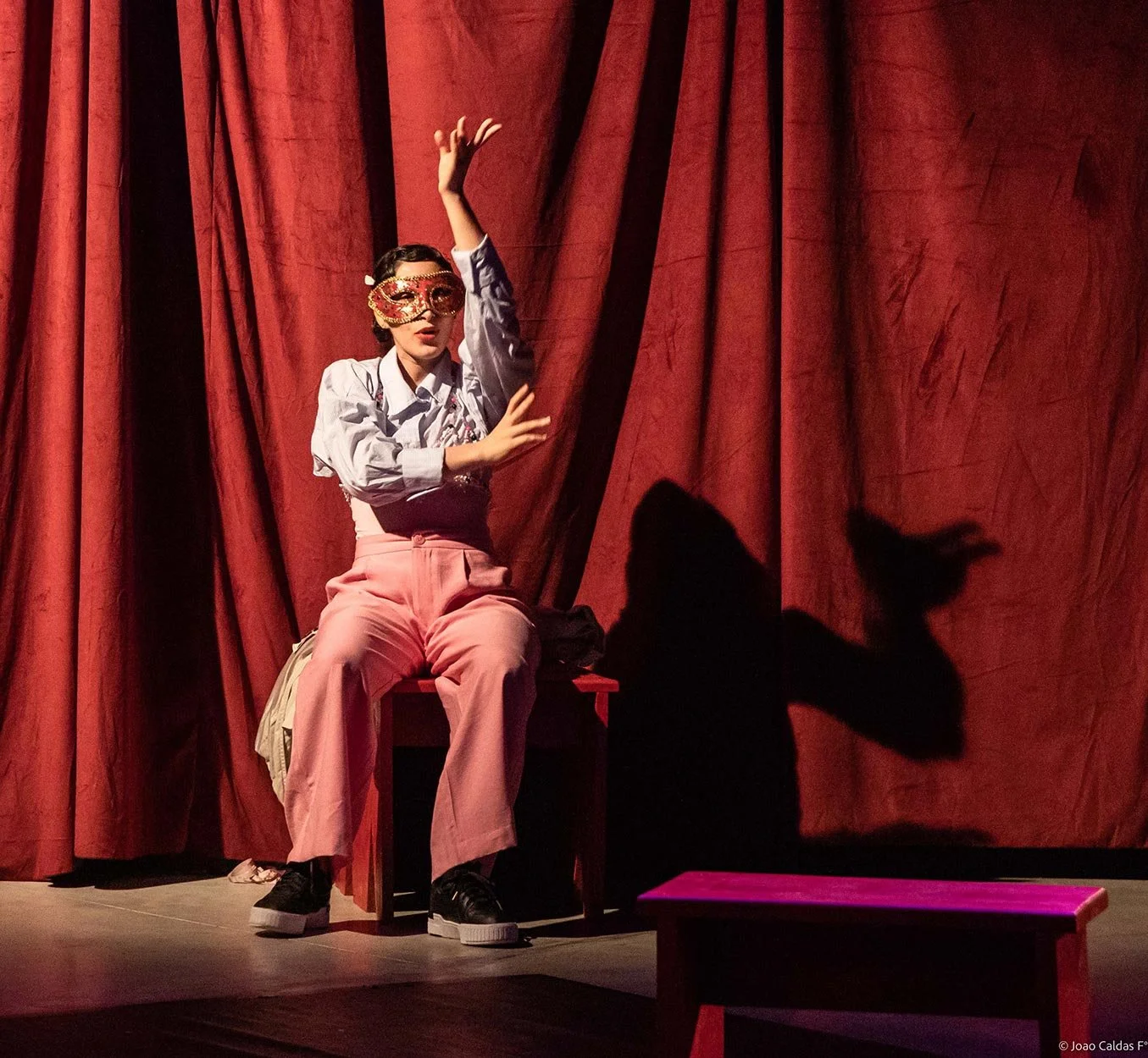 Pessoa sentada em um palco diante de cortinas vermelhas, usando máscara de carnaval, camiseta branca com mangas bufantes, calça rosa claro e tênis pretos, fazendo sinal com as mãos enquanto a sombra é projetada na cortina.
