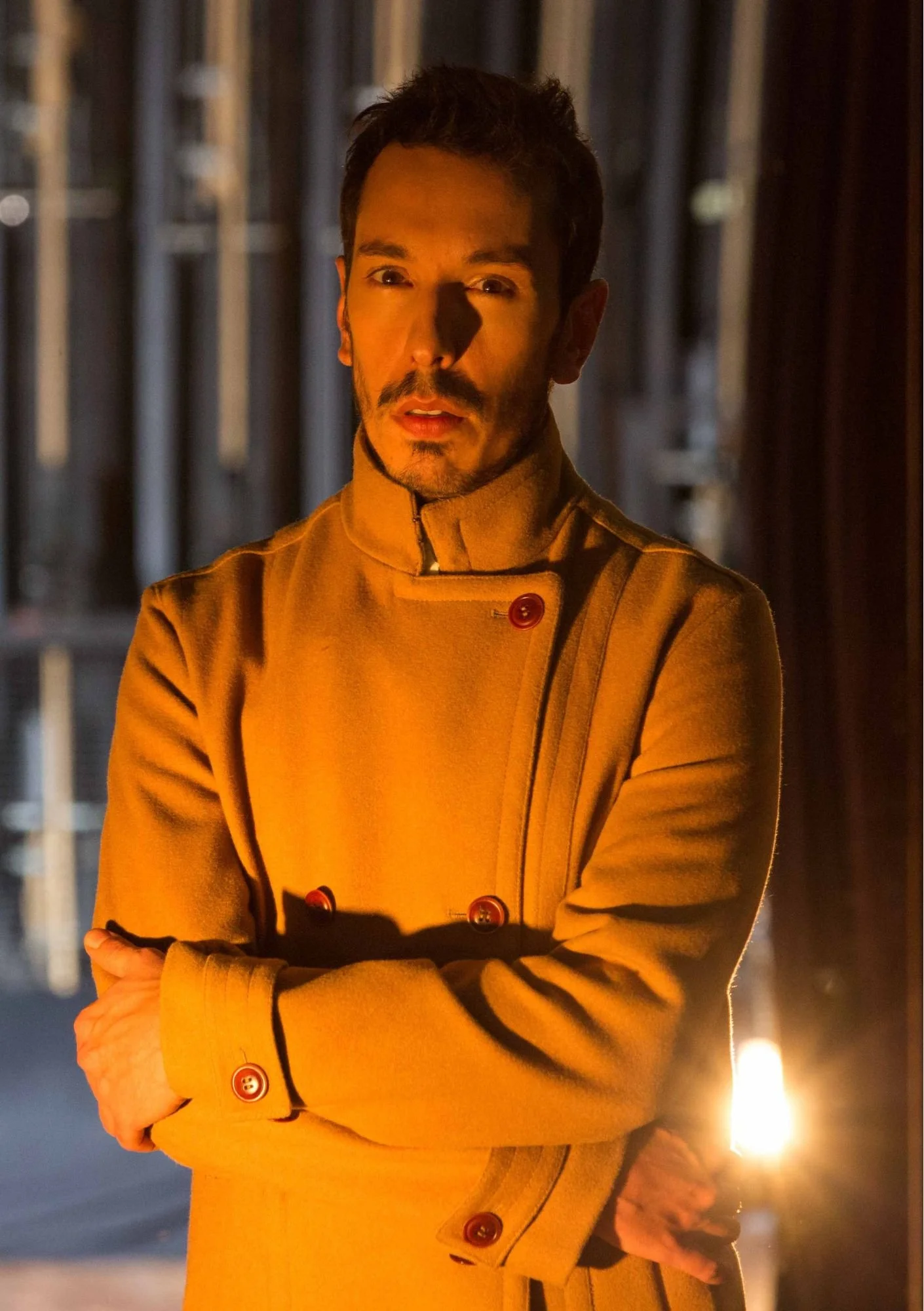 Homem com barba, vestido com casaco amarelo, em ambiente com iluminação quente e fundos de estrutura de madeira.