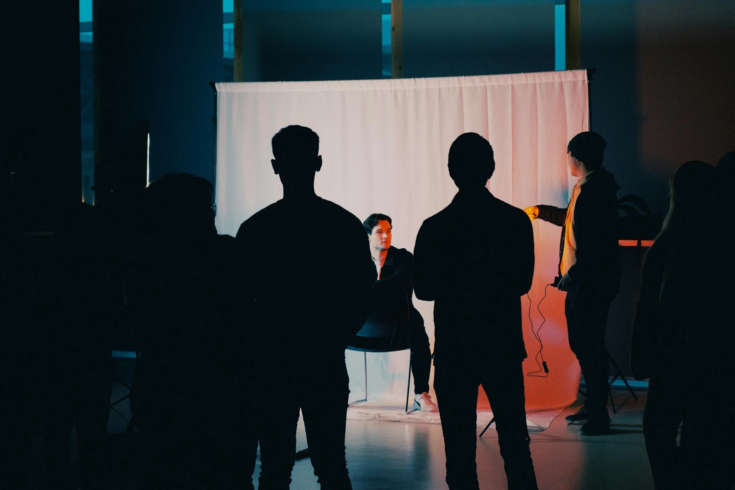 Um grupo de pessoas assistindo a uma gravação com uma pessoa sentada na frente de um fundo branco, enquanto outra pessoa ajusta equipamentos de iluminação ou câmeras, em um ambiente de estúdio com iluminação moderada.