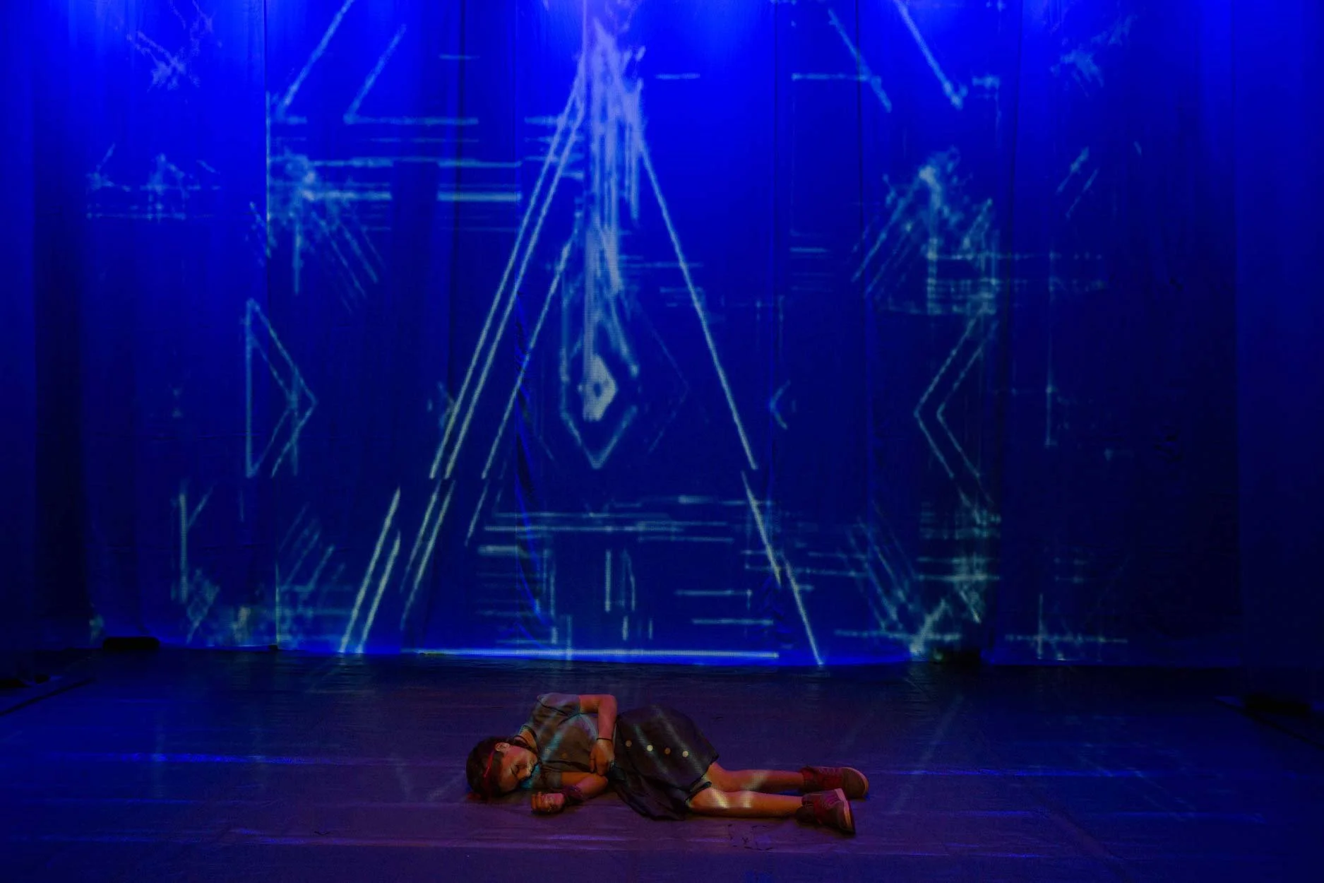 Pessoa deitada no palco, com uma instalação de luzes azuis ao fundo.