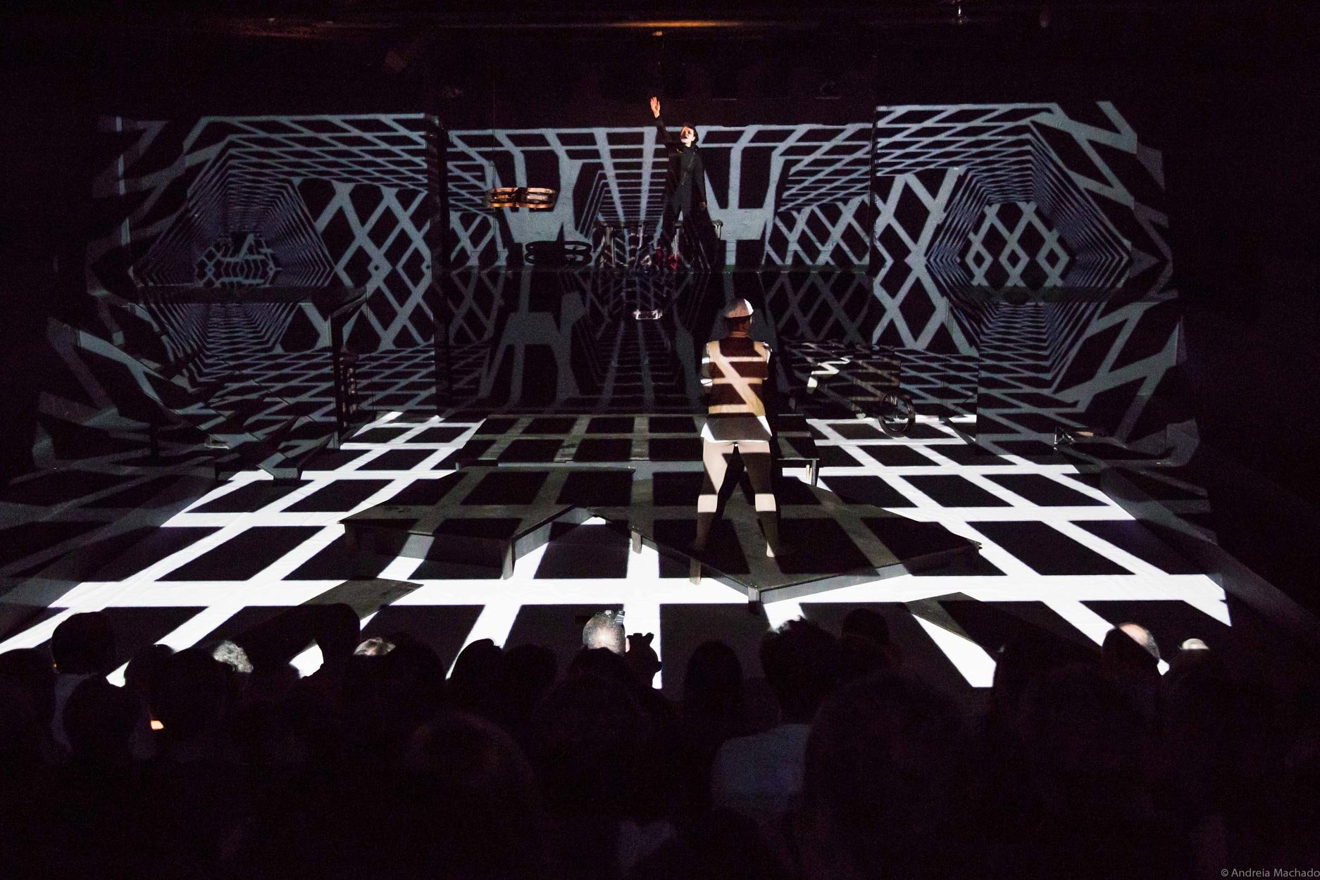 Espetáculo de teatro com projeções visuais tridimensionais em preto e branco no palco, uma mulher com chapéu e roupas de listras na frente, e uma pessoa ao fundo no palco, com público assistindo.