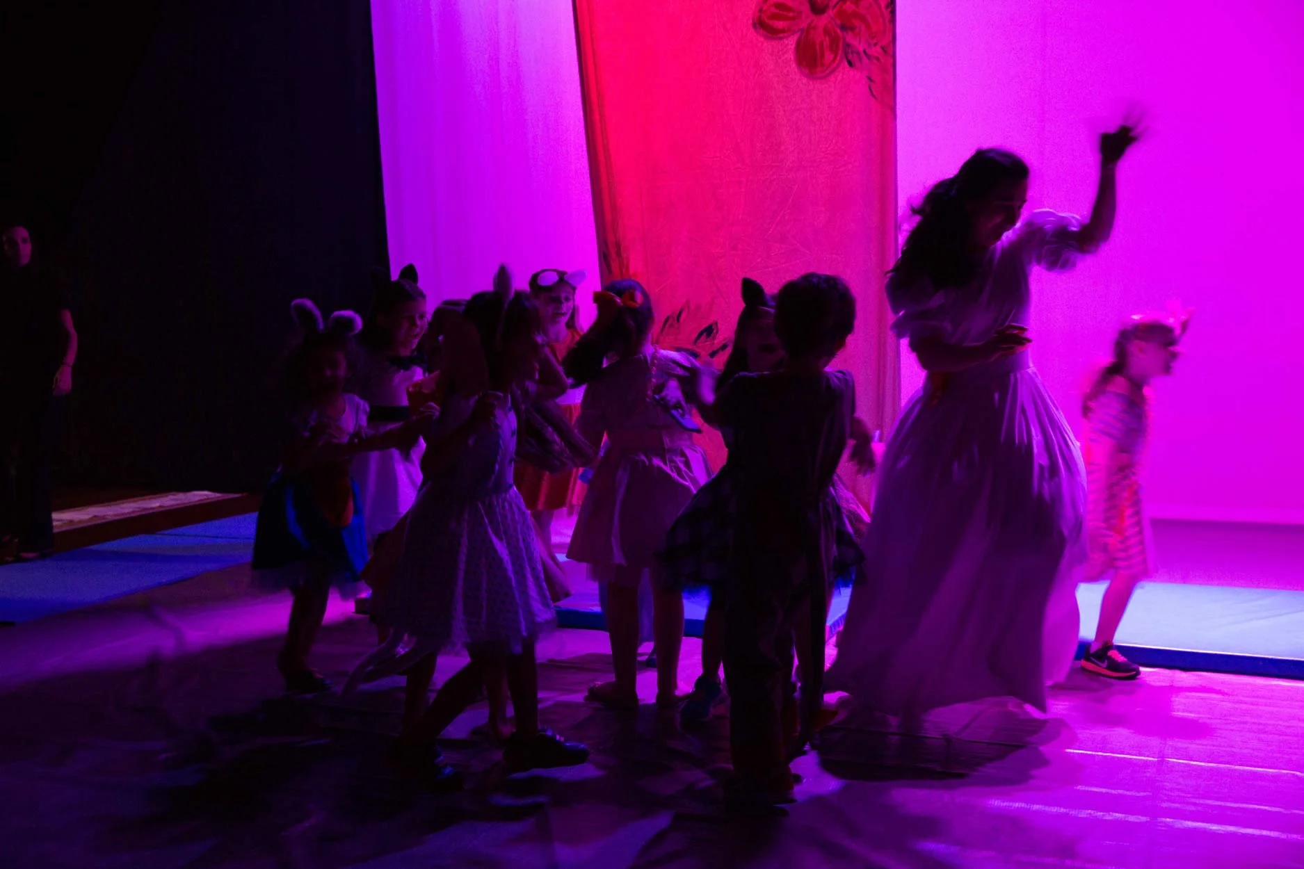 Crianças em uma apresentação de teatro ou dança, usando fantasias de animais em um palco iluminado com luz roxa e rosa.