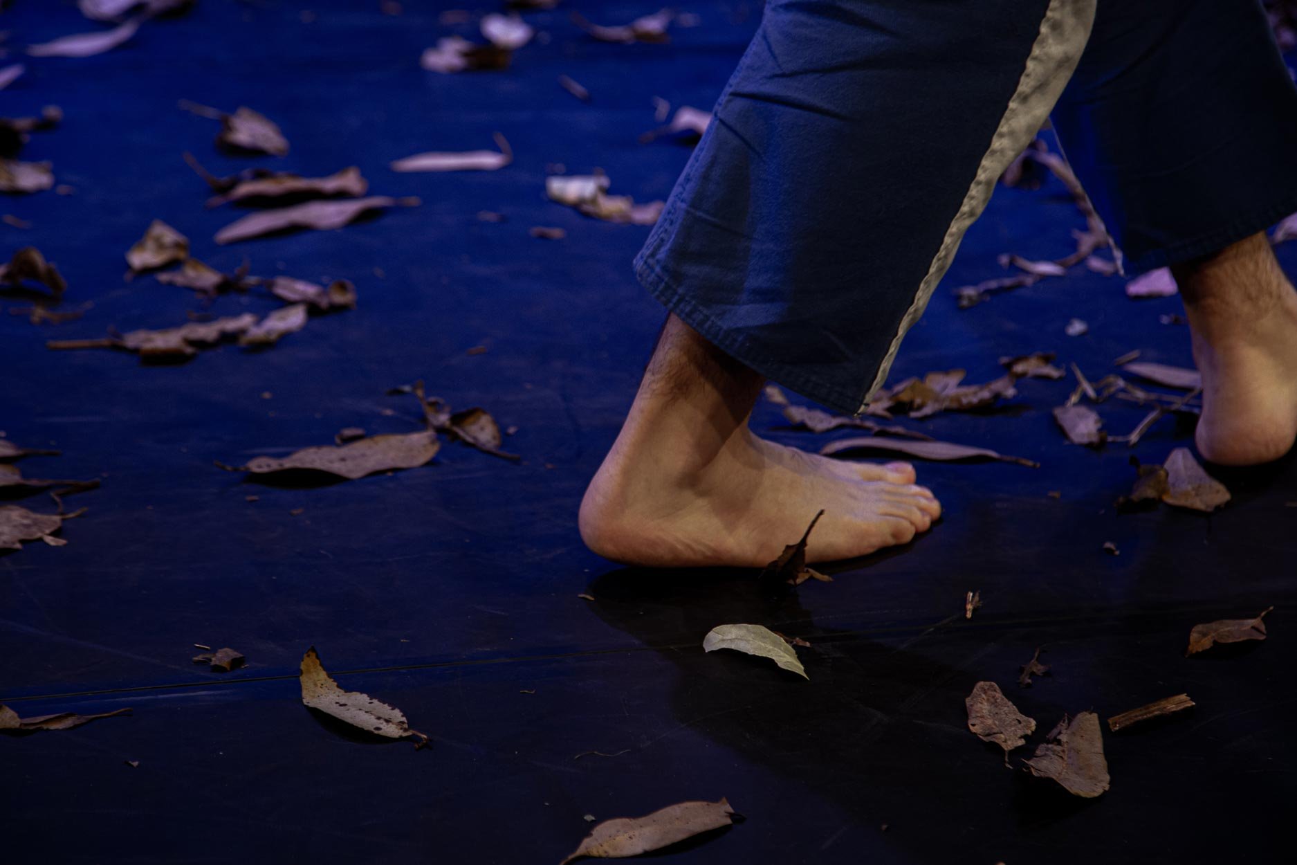 Pés descalços de uma pessoa, deitada ou agachada, com piso preto e folhas secas espalhadas ao redor.
